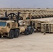A large military staging area or motor pool in an arid desert environment, featuring rows of tan/sand-colored military vehicles lined up in organized formation. In the foreground, an Oshkosh heavy tactical truck with a crane/loader attachment is positioned near stacked military pallets or flatrack containers. At least one uniformed soldier is visible supervising or conducting logistics operations.