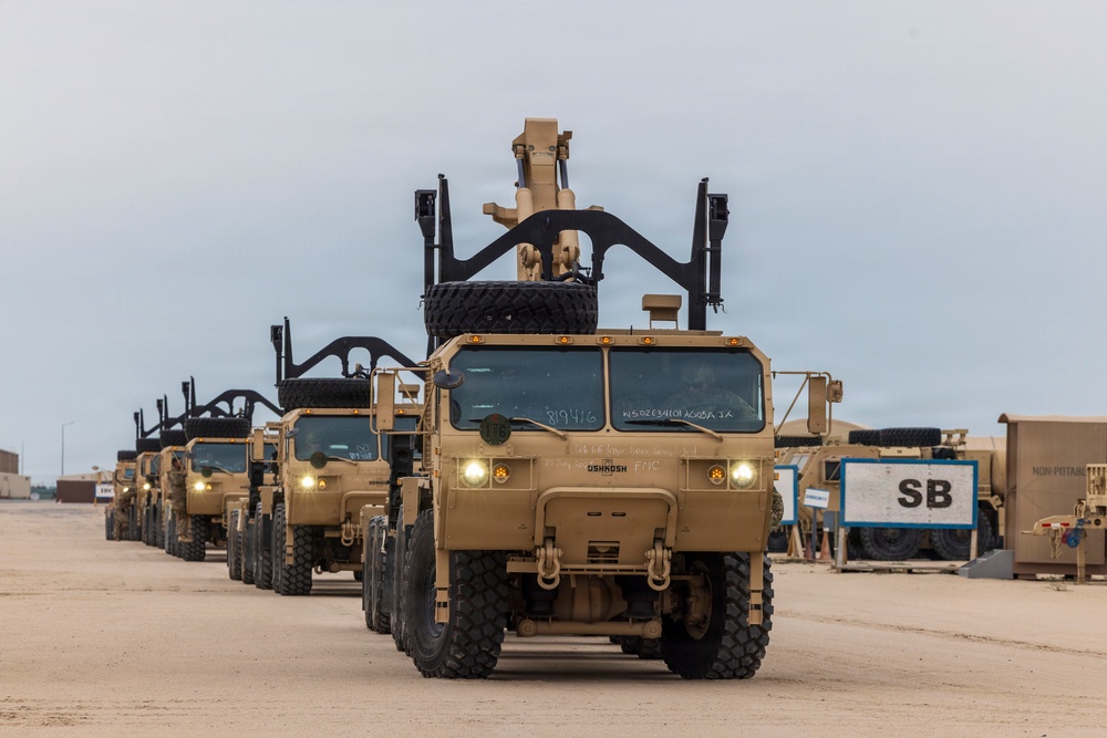 A convoy of tan/desert-colored military trucks, identifiable as Oshkosh-manufactured vehicles, are lined up in formation on a sandy surface. The lead vehicle displays markings including '819416' and 'OSHKOSH' on its front cab, with headlights illuminated. Each vehicle is equipped with a rear-mounted crane or recovery arm apparatus in the raised position.