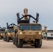 A convoy of tan/desert-colored military trucks, identifiable as Oshkosh-manufactured vehicles, are lined up in formation on a sandy surface. The lead vehicle displays markings including '819416' and 'OSHKOSH' on its front cab, with headlights illuminated. Each vehicle is equipped with a rear-mounted crane or recovery arm apparatus in the raised position.
