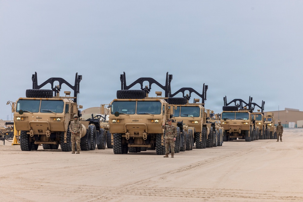 A formation of multiple tan/sand-colored military heavy trucks, consistent with M1070 Heavy Equipment Transporters (HETs) or similar HEMTT-series vehicles, are lined up on a flat sandy surface. Several soldiers in camouflage uniforms are standing beside the vehicles, suggesting a pre-mission inspection, staging, or readiness review. The setting appears to be a forward operating base or staging area in an arid desert environment.