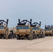 A formation of multiple tan/sand-colored military heavy trucks, consistent with M1070 Heavy Equipment Transporters (HETs) or similar HEMTT-series vehicles, are lined up on a flat sandy surface. Several soldiers in camouflage uniforms are standing beside the vehicles, suggesting a pre-mission inspection, staging, or readiness review. The setting appears to be a forward operating base or staging area in an arid desert environment.