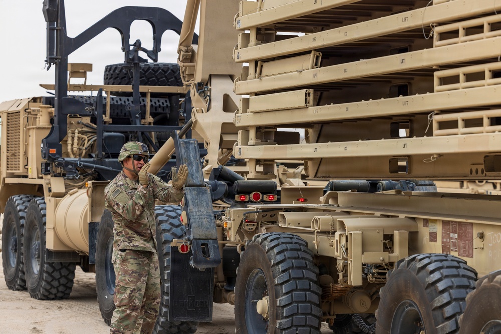 A military service member in OCP (Operational Camouflage Pattern) uniform is working between two large military vehicles in what appears to be an arid or desert environment. The soldier appears to be performing a coupling or equipment check operation between the vehicles. Stacked military equipment panels or ramps are visible loaded on one of the vehicles.