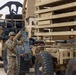 A military service member in OCP (Operational Camouflage Pattern) uniform is working between two large military vehicles in what appears to be an arid or desert environment. The soldier appears to be performing a coupling or equipment check operation between the vehicles. Stacked military equipment panels or ramps are visible loaded on one of the vehicles.