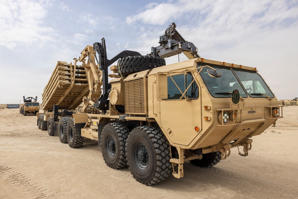 A large military heavy-duty tactical truck, identifiable as an Oshkosh-manufactured vehicle, is parked on a sandy desert terrain. The truck appears to be a Heavy Expanded Mobility Tactical Truck (HEMTT) variant equipped with a crane/loader arm and is hauling a flatbed trailer loaded with military equipment or structural components. The setting suggests a forward operating base or staging area in an arid environment.