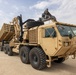 A large military heavy-duty tactical truck, identifiable as an Oshkosh-manufactured vehicle, is parked on a sandy desert terrain. The truck appears to be a Heavy Expanded Mobility Tactical Truck (HEMTT) variant equipped with a crane/loader arm and is hauling a flatbed trailer loaded with military equipment or structural components. The setting suggests a forward operating base or staging area in an arid environment.