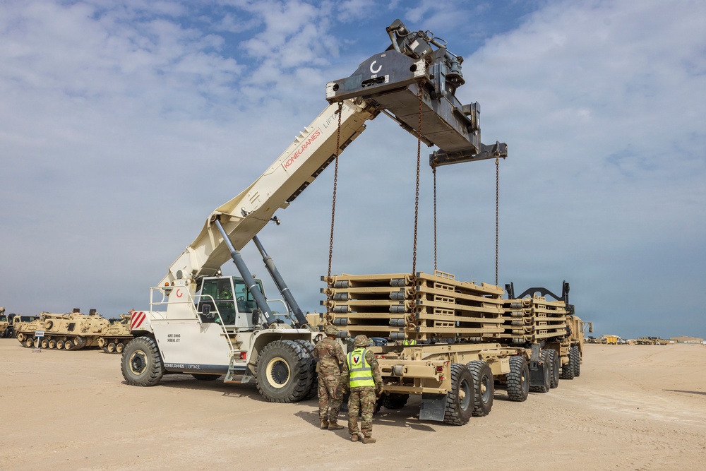 A Konecranes Liftace reach stacker crane is lifting a large stack of military equipment components, likely bridge sections or palletized military hardware, onto a flatbed military trailer in an arid outdoor staging area. Two personnel in military camouflage uniforms, one wearing a high-visibility yellow vest, are supervising the lift operation on the ground. Several military tracked vehicles, including what appears to be an armored recovery vehicle, are visible in the background.