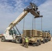 A Konecranes Liftace reach stacker crane is lifting a large stack of military equipment components, likely bridge sections or palletized military hardware, onto a flatbed military trailer in an arid outdoor staging area. Two personnel in military camouflage uniforms, one wearing a high-visibility yellow vest, are supervising the lift operation on the ground. Several military tracked vehicles, including what appears to be an armored recovery vehicle, are visible in the background.