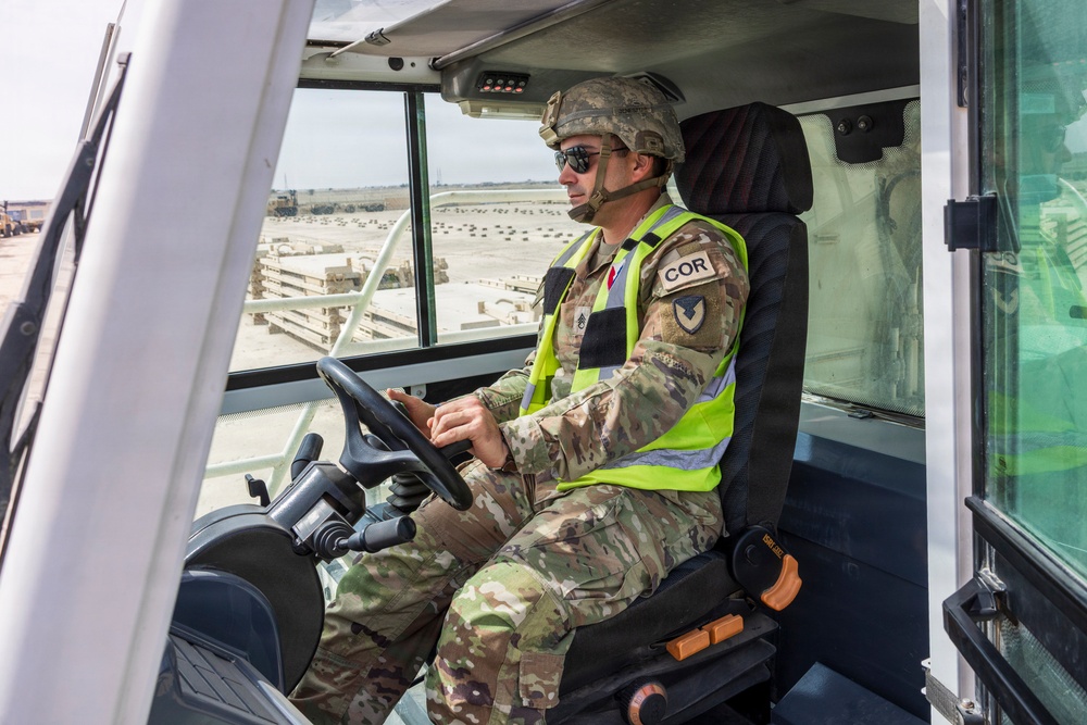 A military personnel member in US Army OCP (Operational Camouflage Pattern) uniform is seated in the cab of a heavy equipment vehicle or forklift. The soldier is wearing a combat helmet, sunglasses, and a high-visibility safety vest with a 'COR' (Contracting Officer's Representative) patch visible on the arm. The background shows what appears to be a military logistics or staging area with pallets and equipment.