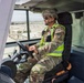 A military personnel member in US Army OCP (Operational Camouflage Pattern) uniform is seated in the cab of a heavy equipment vehicle or forklift. The soldier is wearing a combat helmet, sunglasses, and a high-visibility safety vest with a 'COR' (Contracting Officer's Representative) patch visible on the arm. The background shows what appears to be a military logistics or staging area with pallets and equipment.