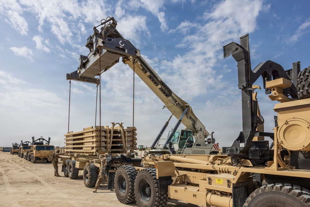 A military logistics operation is underway in an arid, desert-like environment. A large crane or reach stacker is lifting stacked military pallets or equipment frames from a flatbed trailer that is part of a convoy of tan/sand-colored military heavy transport vehicles. At least one uniformed soldier is visible observing or supervising the operation.