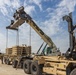 A military logistics operation is underway in an arid, desert-like environment. A large crane or reach stacker is lifting stacked military pallets or equipment frames from a flatbed trailer that is part of a convoy of tan/sand-colored military heavy transport vehicles. At least one uniformed soldier is visible observing or supervising the operation.