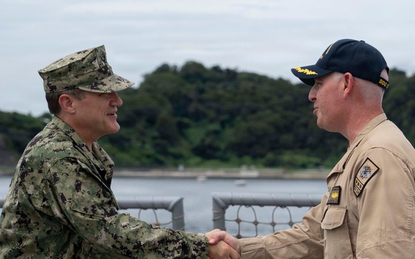Two U.S. Navy personnel are shaking hands on the deck of a naval vessel, likely during an official visit or greeting ceremony. One individual is wearing the Navy Working Uniform (NWU Type III, woodland camouflage) and the other is wearing khaki service uniform with a black command ball cap bearing gold insignia. The background shows a waterway with a tree-covered landmass, suggesting a harbor or port environment.
