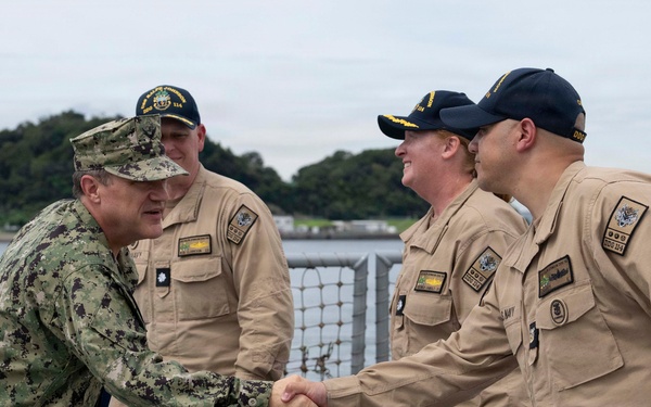 A U.S. Navy officer in digital camouflage (NWU Type III) is shaking hands with personnel wearing tan/khaki Navy Working Uniforms (NWU Type II) aboard what appears to be a naval vessel. The setting appears to be a pier or ship deck with a waterfront and industrial crane visible in the background. The interaction suggests a formal greeting or inspection visit.