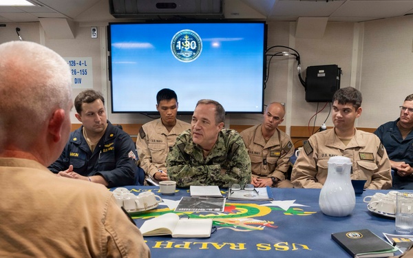 A group of U.S. Navy personnel are gathered around a conference table aboard what appears to be the USS Ralph (visible on the blue tablecloth) for a briefing or meeting. A senior officer in the center appears to be leading the discussion, with a presentation screen displaying a military emblem visible in the background. Personnel are wearing a mix of Navy Working Uniform (NWU) and khaki service uniforms.