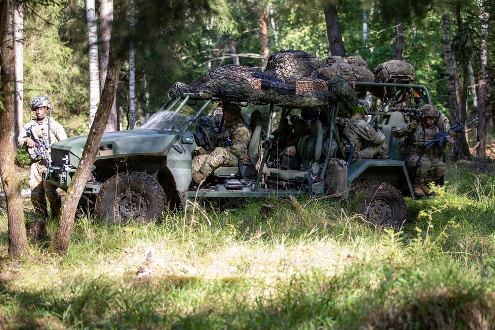 A group of military personnel in full combat gear are positioned around a green tactical off-road vehicle in a forested environment. The soldiers are equipped with weapons, body armor, and camouflage uniforms, suggesting a training exercise or operational deployment. The vehicle is loaded with equipment secured under a cargo net on its roof rack.