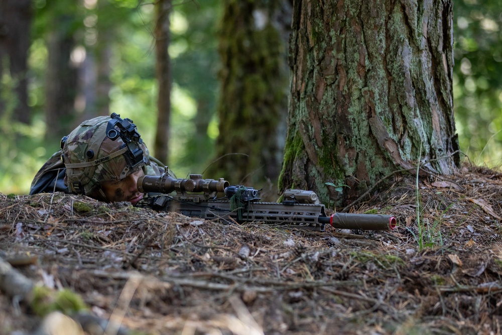 A soldier in full combat gear is lying prone in a forested environment, aiming through a rifle scope. The soldier is positioned behind a large tree trunk, using it as natural cover. The weapon features a red-capped suppressor or barrel protector, suggesting this may be a training exercise.