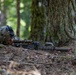 A soldier in full combat gear is lying prone in a forested environment, aiming through a rifle scope. The soldier is positioned behind a large tree trunk, using it as natural cover. The weapon features a red-capped suppressor or barrel protector, suggesting this may be a training exercise.