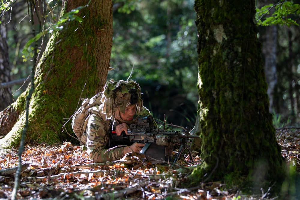 A U.S. Army soldier in full MultiCam camouflage is positioned prone on a forest floor between moss-covered trees, aiming a light machine gun with a bipod deployed. The soldier is wearing a helmet adorned with camouflage netting and vegetation for concealment. The setting appears to be a training exercise in a temperate deciduous forest environment.