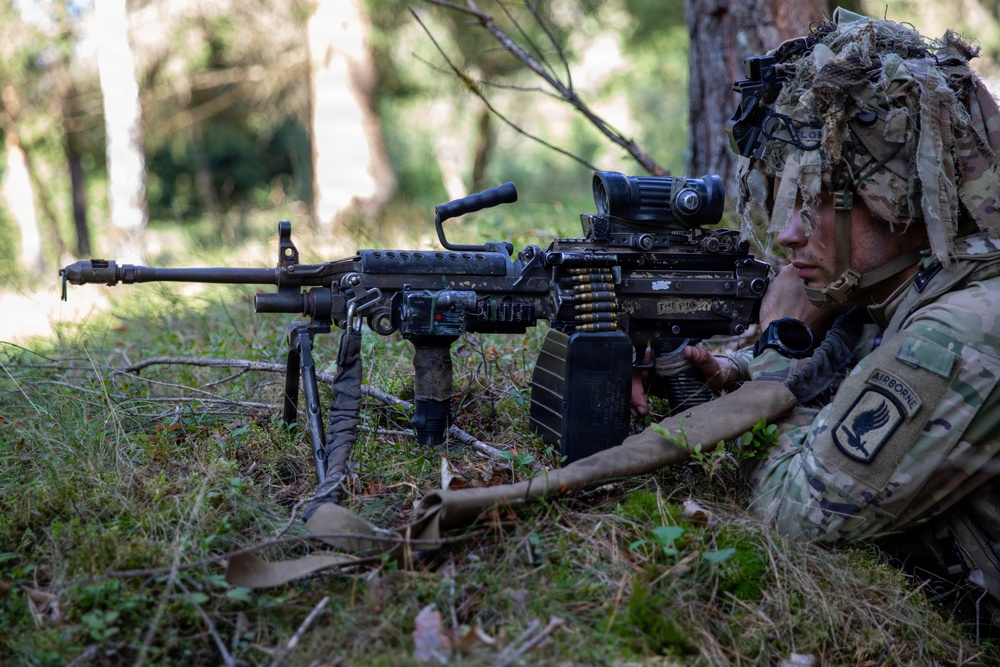 A U.S. Army Airborne soldier is positioned prone in a wooded area, manning a light machine gun on a bipod. The soldier is wearing full camouflage including a ghillie-style helmet cover and has face paint applied. The scene appears to be a field training exercise or tactical operation.