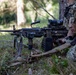 A U.S. Army Airborne soldier is positioned prone in a wooded area, manning a light machine gun on a bipod. The soldier is wearing full camouflage including a ghillie-style helmet cover and has face paint applied. The scene appears to be a field training exercise or tactical operation.