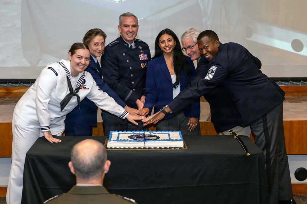 A group of six individuals, including military personnel and civilians, are gathered around a table cutting a decorated ceremonial cake. The central figure is a high-ranking Air Force officer in dress blues with multiple ribbons and medals. The event appears to be a military anniversary or celebration ceremony held in an auditorium setting.