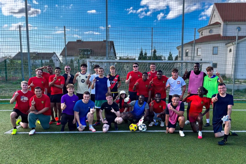 A large group of approximately 25 young men pose for a team photo on an outdoor artificial turf football/soccer pitch. Most participants are making a 'number one' finger gesture toward the camera, suggesting they are celebrating a victory or simply posing enthusiastically. The setting appears to be a recreational or amateur football facility in what looks like a Central/Eastern European town.