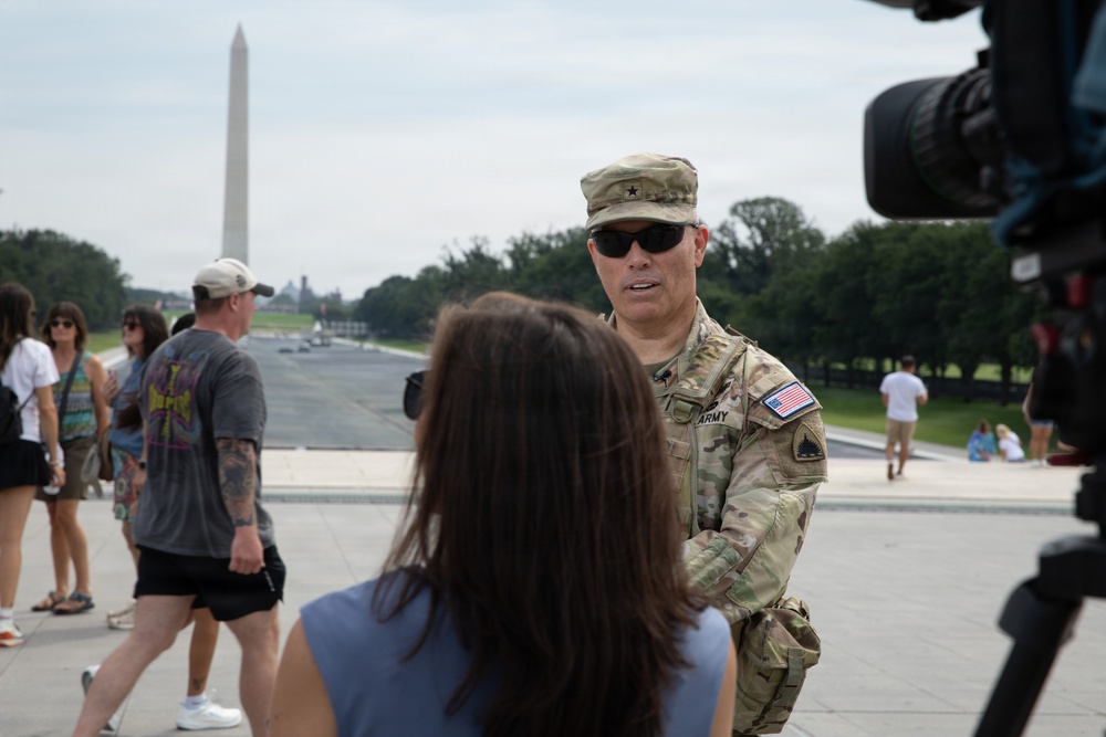 A U.S. Army officer wearing a one-star general rank insignia on his patrol cap is being interviewed by a reporter near the National Mall in Washington, D.C. A professional video camera is visible on the right side of the frame. The Washington Monument and the Reflecting Pool are clearly visible in the background.