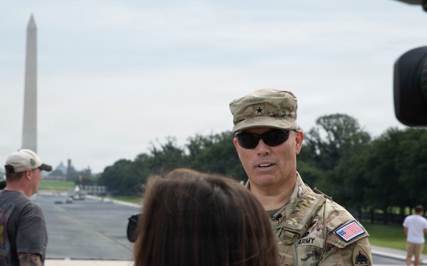 A U.S. Army officer wearing a one-star general rank insignia on his patrol cap is being interviewed by a reporter near the National Mall in Washington, D.C. A professional video camera is visible on the right side of the frame. The Washington Monument and the Reflecting Pool are clearly visible in the background.