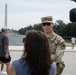 A U.S. Army officer wearing a one-star general rank insignia on his patrol cap is being interviewed by a reporter near the National Mall in Washington, D.C. A professional video camera is visible on the right side of the frame. The Washington Monument and the Reflecting Pool are clearly visible in the background.