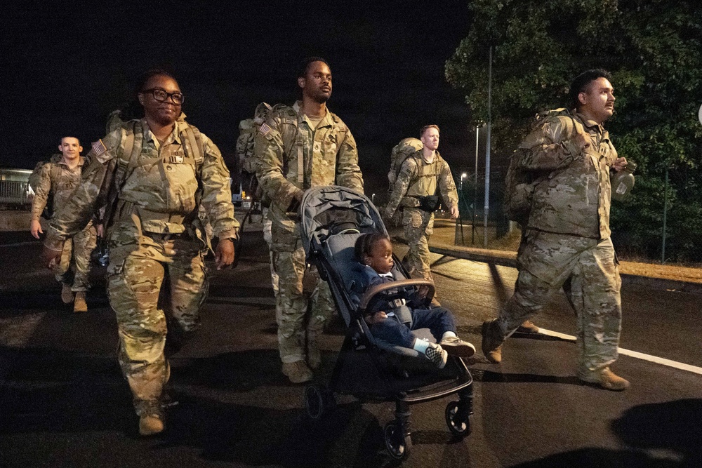 A group of military personnel in camouflage uniforms are walking together at night, carrying large rucksacks on their backs. One soldier is pushing a stroller with a young child seated in it. The scene appears to be on a paved road or parking area, likely at or near a military installation.