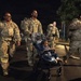 A group of military personnel in camouflage uniforms are walking together at night, carrying large rucksacks on their backs. One soldier is pushing a stroller with a young child seated in it. The scene appears to be on a paved road or parking area, likely at or near a military installation.