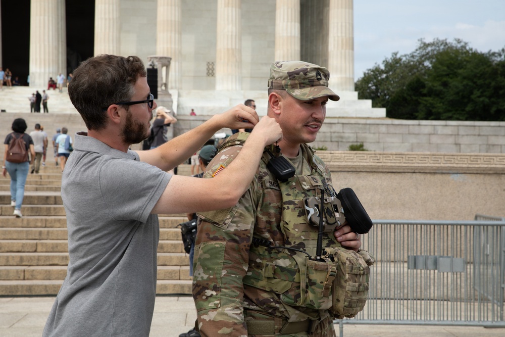 A civilian in a gray polo shirt appears to be adjusting a microphone or earpiece on a uniformed U.S. Army soldier in MultiCam (OCP) camouflage. The setting is the Lincoln Memorial in Washington, D.C., with its distinctive columns and steps visible in the background. The interaction suggests a media or interview setup, possibly for a news broadcast or documentary.