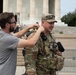 A civilian in a gray polo shirt appears to be adjusting a microphone or earpiece on a uniformed U.S. Army soldier in MultiCam (OCP) camouflage. The setting is the Lincoln Memorial in Washington, D.C., with its distinctive columns and steps visible in the background. The interaction suggests a media or interview setup, possibly for a news broadcast or documentary.