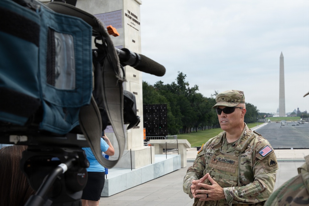 A U.S. Army soldier, identified by name tape as 'Blanchard,' is being interviewed on camera near the National Mall in Washington, D.C. The Washington Monument is clearly visible in the background, along with the reflecting pool area. The soldier is in full camouflage uniform with a general officer star visible on his cap.