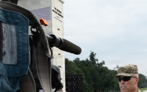 A U.S. Army soldier, identified by name tape as 'Blanchard,' is being interviewed on camera near the National Mall in Washington, D.C. The Washington Monument is clearly visible in the background, along with the reflecting pool area. The soldier is in full camouflage uniform with a general officer star visible on his cap.