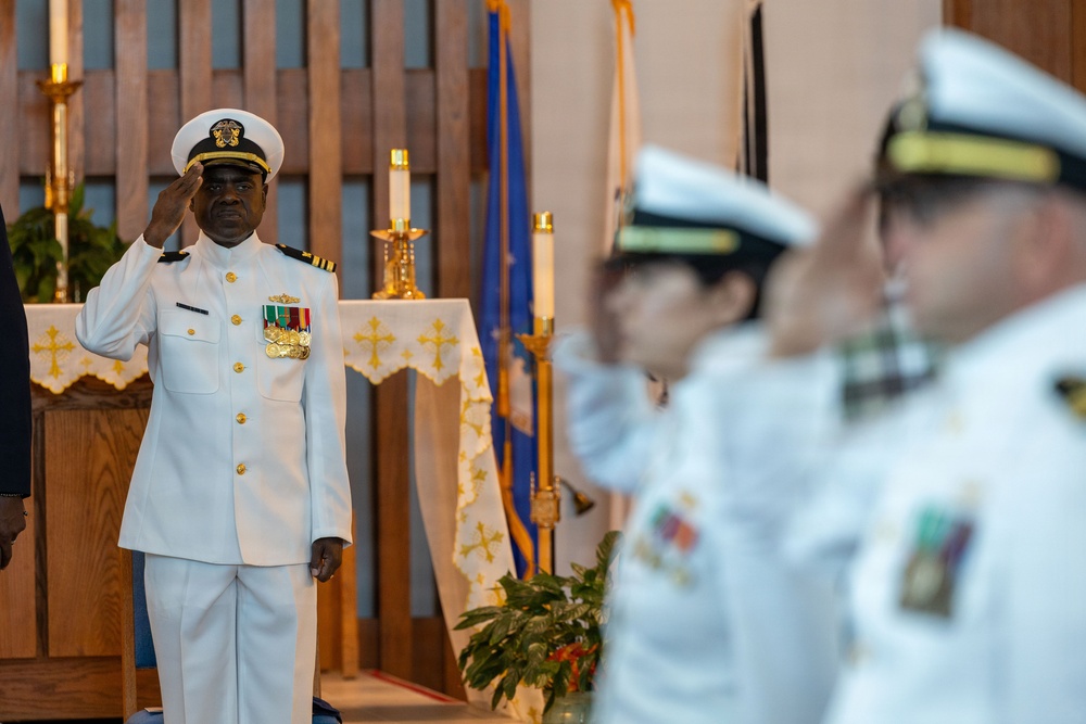 A military ceremony is taking place inside what appears to be a chapel or religious venue. A naval officer in white dress uniform is rendering a salute in the foreground, with additional uniformed personnel visible in the background. The setting includes religious iconography such as crosses on altar cloth and candles, suggesting a formal military religious or ceremonial event.