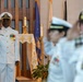 A military ceremony is taking place inside what appears to be a chapel or religious venue. A naval officer in white dress uniform is rendering a salute in the foreground, with additional uniformed personnel visible in the background. The setting includes religious iconography such as crosses on altar cloth and candles, suggesting a formal military religious or ceremonial event.
