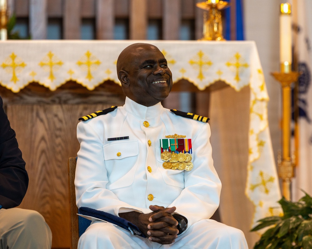 A military officer in a white dress uniform is seated in what appears to be a church or chapel setting, smiling broadly. He is wearing multiple medals, ribbons, and gold insignia on his uniform, with gold epaulettes indicating a senior rank. The background features a white altar cloth decorated with gold crosses and a lit candle.