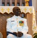 A military officer in a white dress uniform is seated in what appears to be a church or chapel setting, smiling broadly. He is wearing multiple medals, ribbons, and gold insignia on his uniform, with gold epaulettes indicating a senior rank. The background features a white altar cloth decorated with gold crosses and a lit candle.