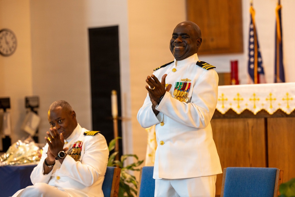 Two men in white U.S. Navy dress uniforms are photographed inside what appears to be a church or chapel setting. The standing officer is smiling broadly and clapping, while the seated officer is also applauding. Both wear multiple service medals and gold insignia consistent with senior naval officer ranks.