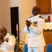 Two men in white U.S. Navy dress uniforms are photographed inside what appears to be a church or chapel setting. The standing officer is smiling broadly and clapping, while the seated officer is also applauding. Both wear multiple service medals and gold insignia consistent with senior naval officer ranks.