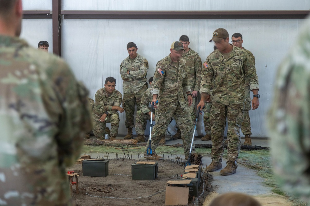 A group of U.S. Army soldiers in OCP (Operational Camouflage Pattern) uniforms are gathered inside a covered training facility around what appears to be a terrain model or sand table exercise. Two soldiers in the center are actively manipulating the model using long-handled tools, while others observe from the background. The scene depicts a military planning or training exercise with simulated terrain features and ammunition cans used as props or markers.