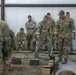 A group of U.S. Army soldiers in OCP (Operational Camouflage Pattern) uniforms are gathered inside a covered training facility around what appears to be a terrain model or sand table exercise. Two soldiers in the center are actively manipulating the model using long-handled tools, while others observe from the background. The scene depicts a military planning or training exercise with simulated terrain features and ammunition cans used as props or markers.
