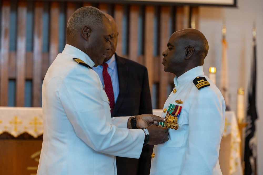A formal military ceremony is taking place in what appears to be a chapel or ceremonial hall. A senior naval officer in a white dress uniform is pinning a medal or decoration onto the chest of another naval officer, also in white dress uniform. A civilian in a dark suit observes the ceremony in the background.