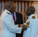 A formal military ceremony is taking place in what appears to be a chapel or ceremonial hall. A senior naval officer in a white dress uniform is pinning a medal or decoration onto the chest of another naval officer, also in white dress uniform. A civilian in a dark suit observes the ceremony in the background.