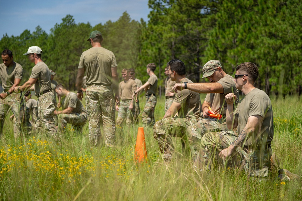 A group of military personnel in camouflage and olive drab uniforms are conducting an outdoor field training exercise in a grassy clearing surrounded by pine trees. One individual in the foreground appears to be instructing or directing another soldier, while other personnel are engaged in various tasks in the background. An orange traffic cone is visible in the mid-ground, suggesting a marked training area.