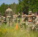 A group of military personnel in camouflage and olive drab uniforms are conducting an outdoor field training exercise in a grassy clearing surrounded by pine trees. One individual in the foreground appears to be instructing or directing another soldier, while other personnel are engaged in various tasks in the background. An orange traffic cone is visible in the mid-ground, suggesting a marked training area.
