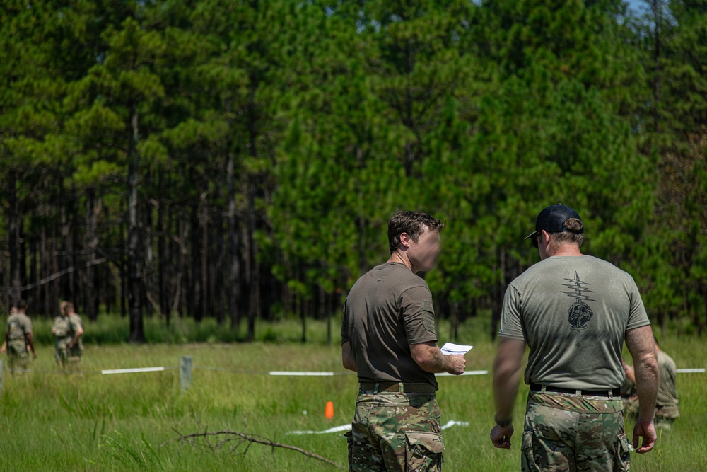 Two individuals in military-style camouflage pants and olive/gray t-shirts stand in a grassy field reviewing a document, with a forested background of pine trees. White tape lines are visible on the ground, suggesting a marked course or range area. Additional personnel in full camouflage gear are visible in the background.