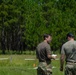 Two individuals in military-style camouflage pants and olive/gray t-shirts stand in a grassy field reviewing a document, with a forested background of pine trees. White tape lines are visible on the ground, suggesting a marked course or range area. Additional personnel in full camouflage gear are visible in the background.