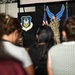 A female military service member in OCP (Operational Camouflage Pattern) uniform is addressing an audience from a stage, holding a microphone and gesturing with one finger raised. The setting appears to be an Air Force Reserve Command event, as indicated by the visible unit insignia and the large Air Force symbol displayed on the backdrop. Several audience members, predominantly women, are visible in the foreground watching the presentation.
